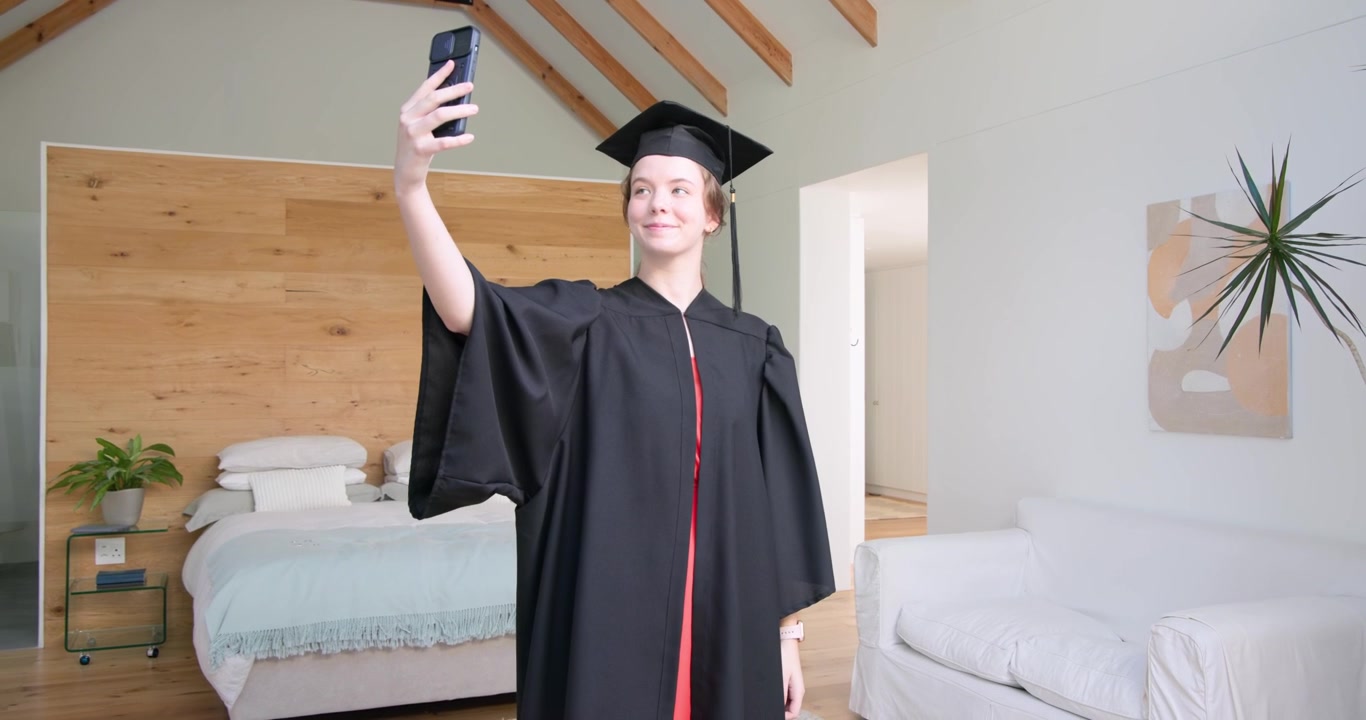 Studentessa che registra un selfie di laurea a casa con il telefono, tocco e toga, leggendo un messaggio e sorridendo.