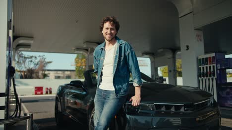 A-young-brunette-guy-in-a-denim-jacket-poses-and-looks-at-the-camera,-leaning-on-his-glossy-dark-gray-convertible