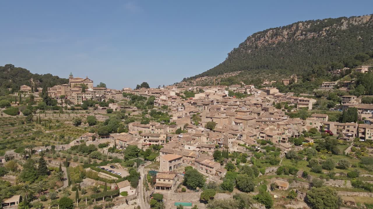 An aerial video of a traditional Mediterranean village surrounded by lush green hills and a rugged mountain backdrop. The tightly packed stone houses with terracotta roofs.