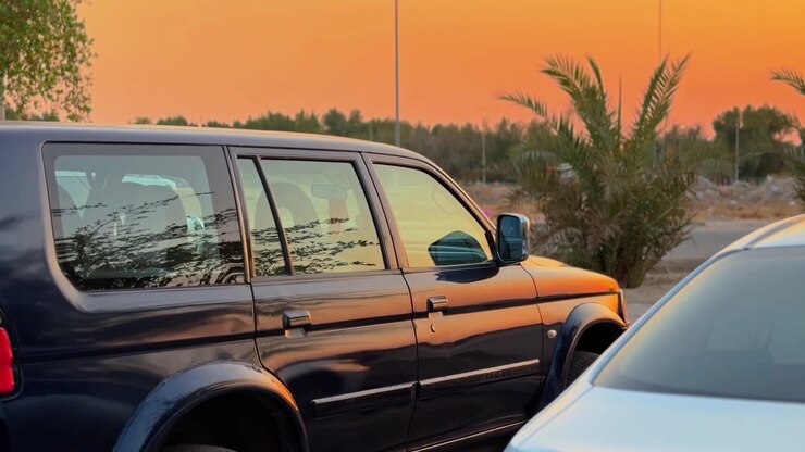 Un SUV scuro e un'auto argentata parcheggiati in un piazzale all'aperto durante un vibrante tramonto arancione. La calda luce dell'ora d'oro si riflette sui finestrini dei veicoli, circondati da palme tropicali in un ambiente tranquillo