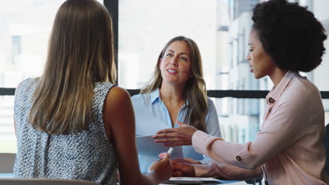 female multi-cultural business team meet around boardroom table with laptops discussing documents