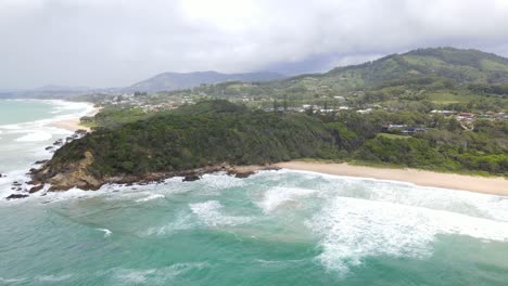 Paisaje-Idílico-De-Olas-Oceánicas-Que-Se-Lavan-En-La-Playa-De-Arena-Con-Vegetación-Exuberante-Y-Montañas-En-La-Playa-De-Zafiro,-Puerto-De-Coffs,-Nsw,-Australia---Toma-Aérea-De-Drones