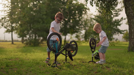 two young siblings are playing outdoors in a grassy field, rotating the wheels of their bicycles that are turned upside down