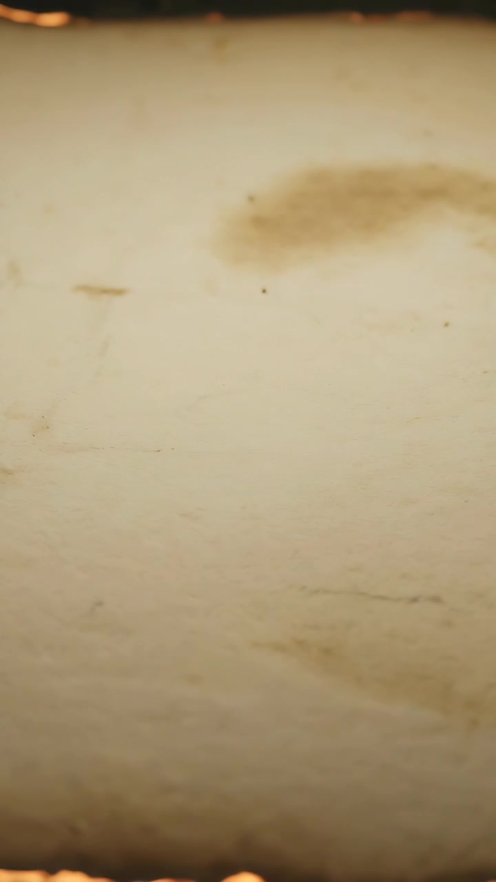 Video verticale: Braci ardenti che causano l'annerimento del bordo di una pergamena invecchiata su una superficie scura, con bordo carbonizzato.