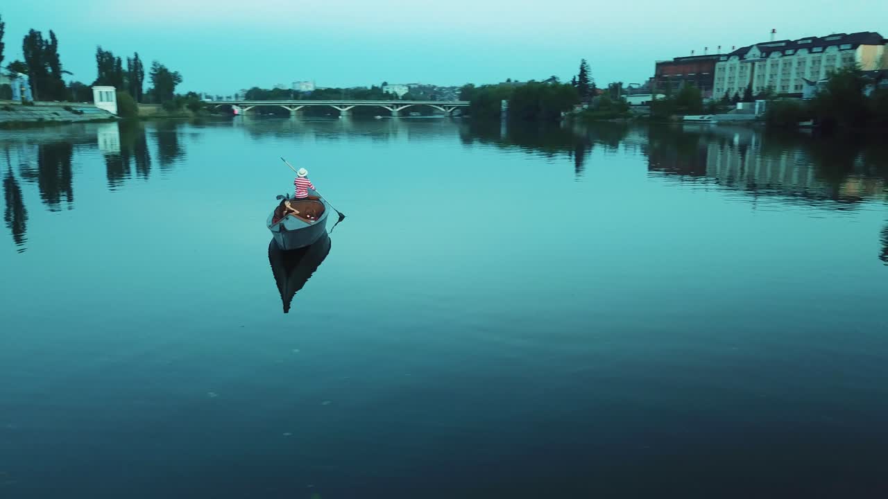 Romantic view of a beautiful woman in a boat and a man in hat rowing on the evening. Calm blue water of a river and gondola with a female relaxing at a summer night.