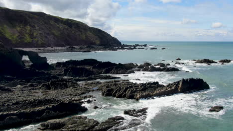 Toma-Aérea-De-Las-Olas-En-El-Mar-Y-Las-Rocas-Y-La-Ladera-En-La-Playa-Costera-De-Spekes-Mill-En-Devon