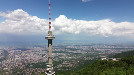 Toma-Aérea,-Acercándose-A-Una-Torre-De-Televisión,-En-Lo-Alto-De-Una-Montaña,-Con-Una-Ciudad-Y-Un-Cielo-Azul-Con-Nubes-Blancas-Y-Esponjosas-En-El-Fondo