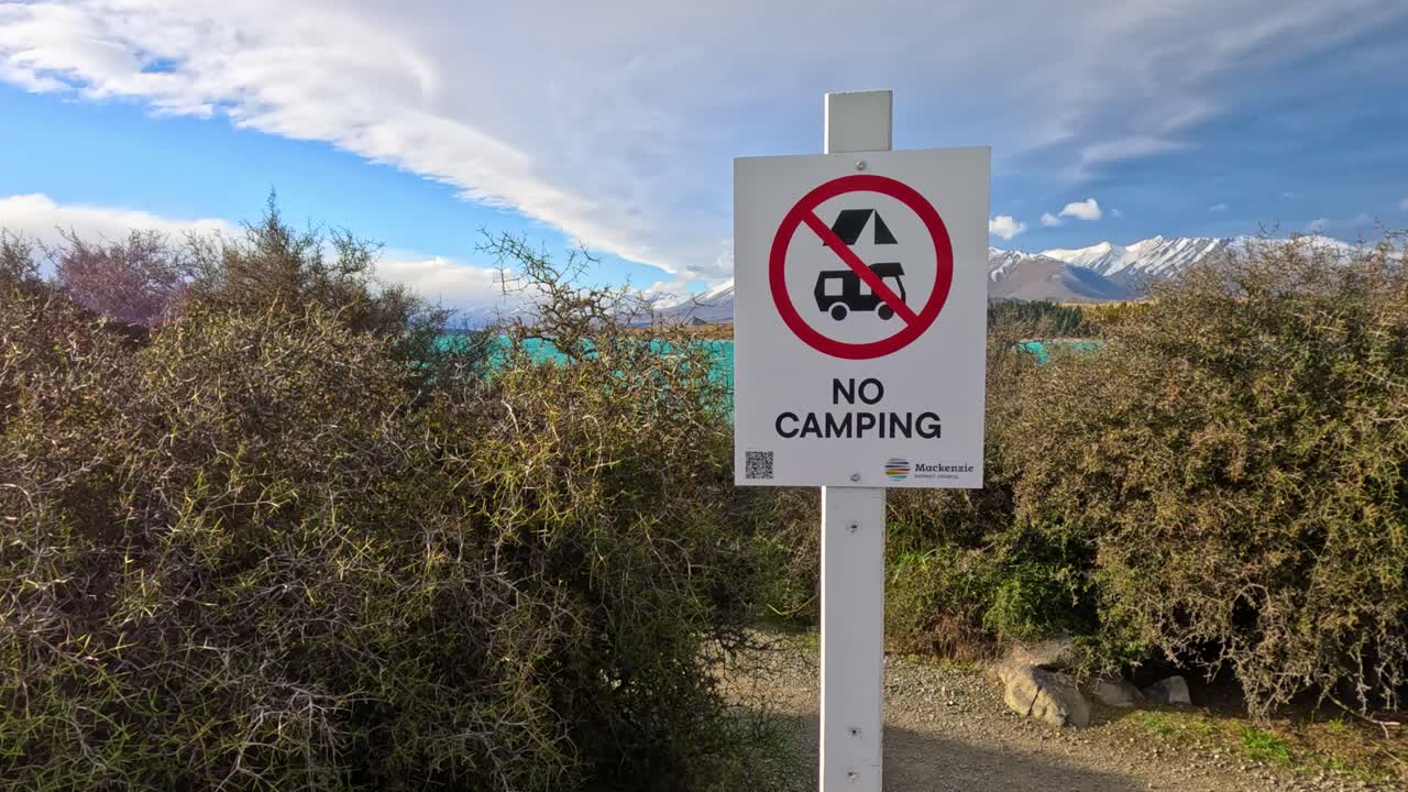 A stationary camera captures a 'No Camping' warning sign in front of dense bushes, with mountains and blue sky in the background. Natural daylight, minimal camera movement