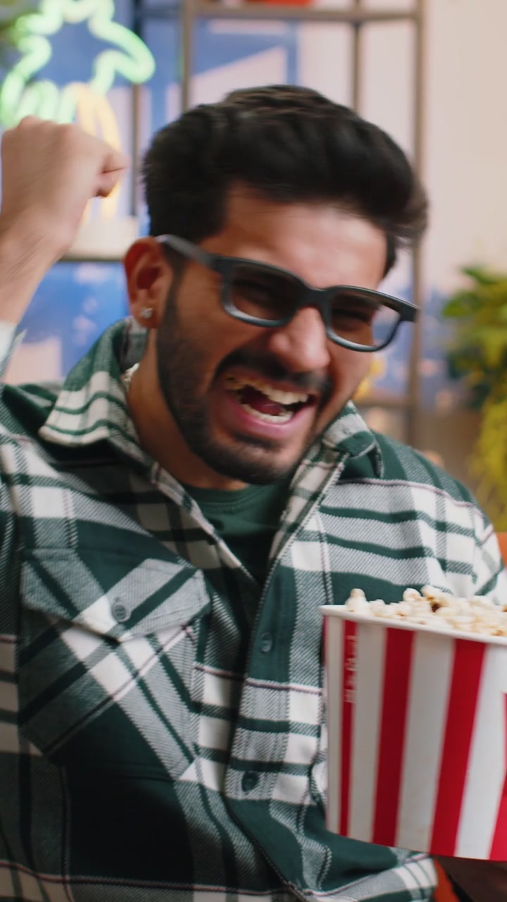 Indian man sits on sofa eating popcorn and watching interesting tv serial sport game online at home