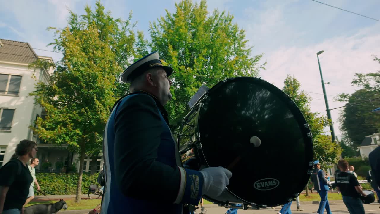 A brass band member in uniform plays a large drum during a parade through the streets of Oosterbeek, Netherlands. The event shows cultural pride and musical tradition