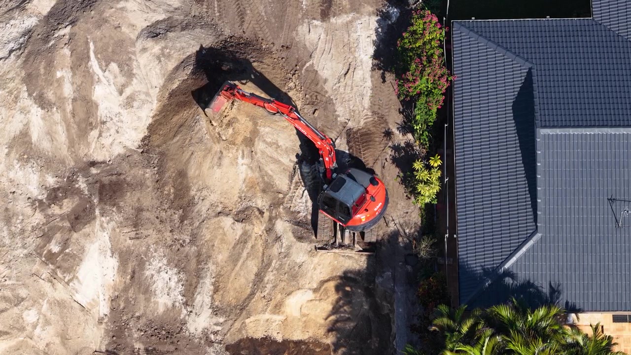 Droneoptagelse ovenfra af en rød gravemaskine, der flytter jord under en renovering af baghaven.