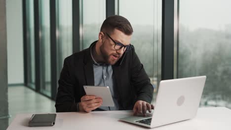 Ein-Beschäftigter-Mann,-Der-Im-Büro-Sitzt,-Beantwortet-Einen-Anruf-Am-Telefon,-Während-Er-Gleichzeitig-An-Einem-Laptop-Und-Einem-Tablet-Arbeitet