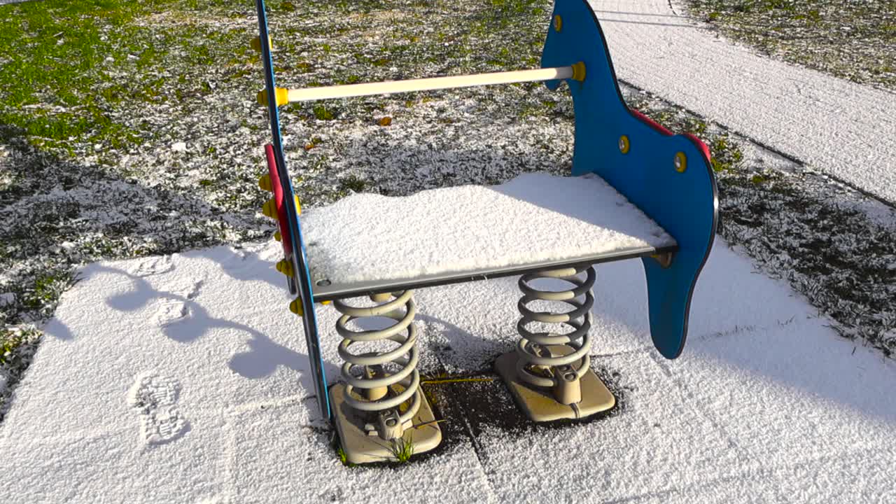 First white snow covered spring rider or bouncer with horses at a sunny winter playground with green grass still visible. The attraction is shaking and moving back and fourth in slow motion