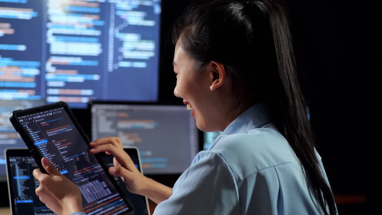 Close Up Back View Of Asian Female Programmer Looking At Database On Tablet While Writing Code By A Laptop Using Multiple Monitors Showing Database On Desktops In The Office