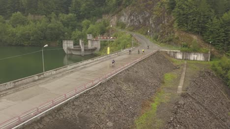 Vista-Aérea-De-Un-Grupo-De-Ciclistas-En-Bicicleta-En-La-Presa-En-El-Lago-Colibita-En-Rumania-Durante-La-Carrera-Tura-Cu-Copaci