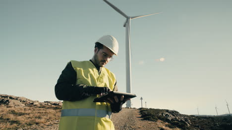 a professional focused engineer in a white helmet and reflective vest uses technology to audit wind turbines in a field of renewable energy generators on a sunny day, promoting eco-friendly energy