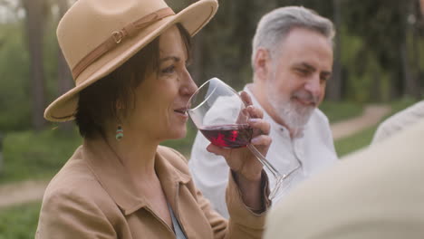 mujer de mediana edad bebiendo vino tinto mientras se sienta a la mesa durante una fiesta al aire libre en el parque