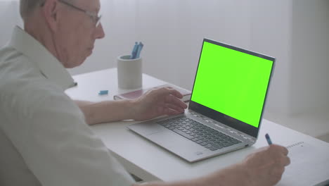 old-man-is-looking-at-screen-of-laptop-and-writing-notes-in-notebook-chroma-key-technology-for-post-production