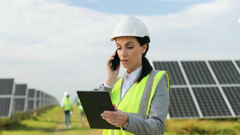 portrait of beautiful female engineer in protective helmet and uniform talking on the phone on solar plantation