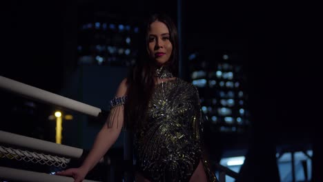 joven latina se para en una escalera por la noche con un vestido brillante con luces de la ciudad en el fondo