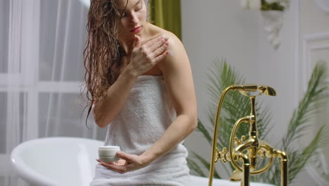 mujer aplicando crema en su piel en el baño mientras está sentada en la bañera