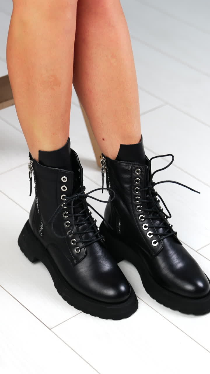 Lady wearing beige coat sits on the stool shows black boots with laces. Beautiful stylish leather boots presentation. White backdrop. Vertical video