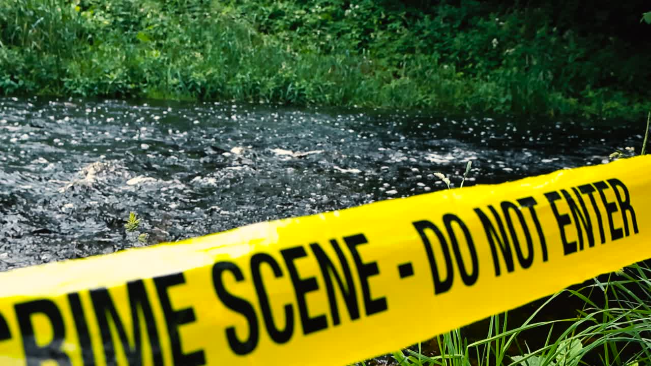 Close up or closeup of a vibrant yellow colored Crime Scene Do Not Enter police caution tape ribbon pulled in front of a fast flowing river shoreline during a cloudy day in nature. focus changes slow
