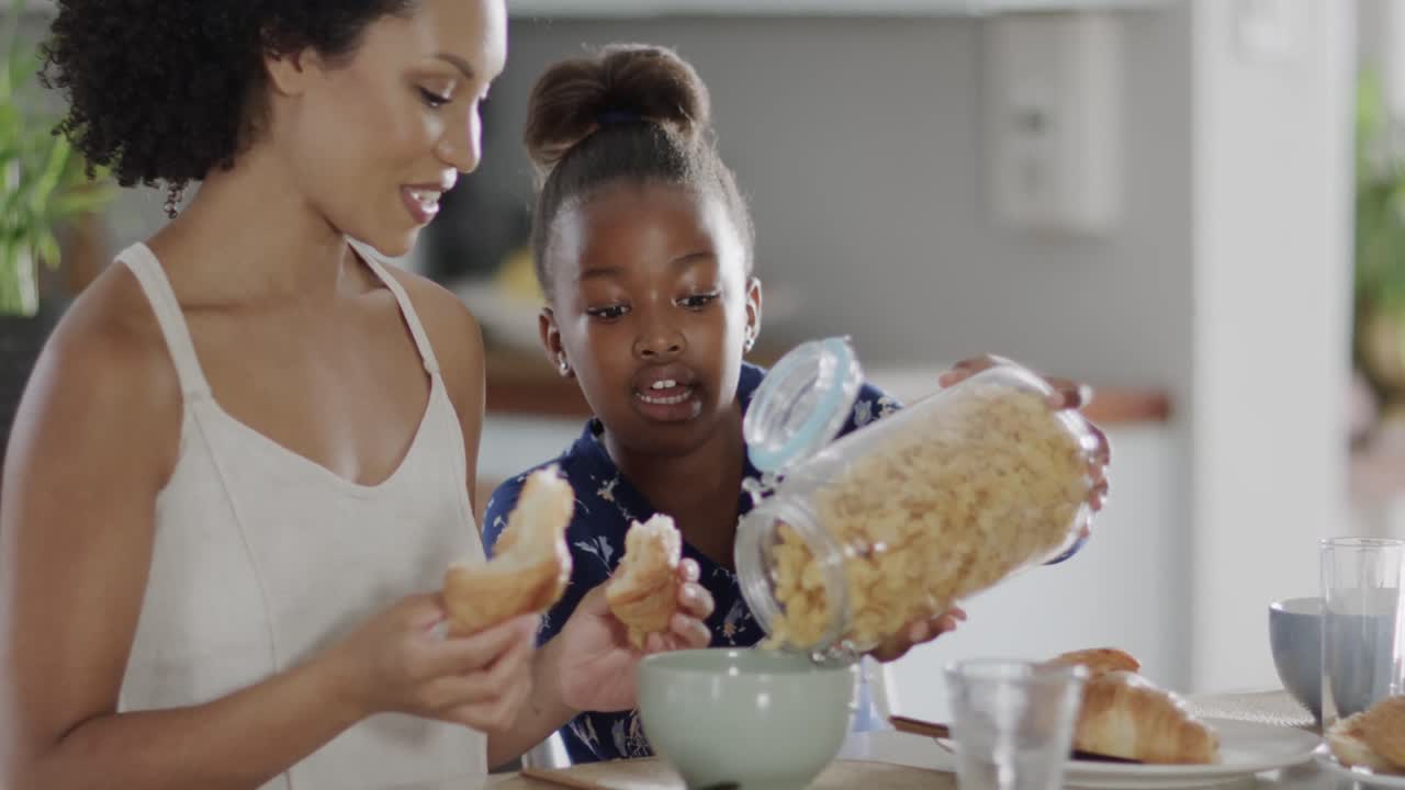 Happy african american mother and daughter eating breakfast in kitchen, slow motion