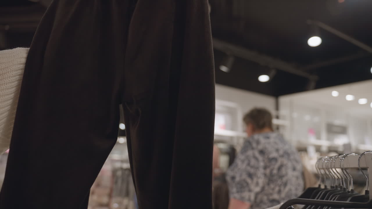 Closeup shot of hand lifting trackies in clothing store, manicured nails feeling fabric on hanger while shoppers browse racks in background, retail scene showing tactile interaction with garment