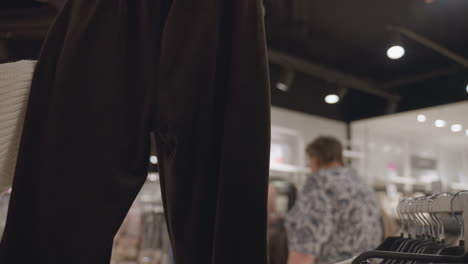 closeup shot of hand lifting trackies in clothing store, manicured nails feeling fabric on hanger while shoppers browse racks in background, retail scene showing tactile interaction with garment