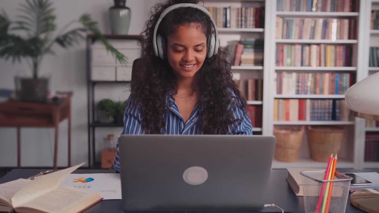Young indian woman putting on headphones and working on laptop at home office
