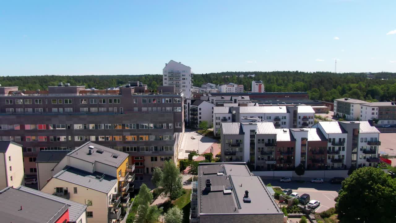 Ekspansivt droneopptak som stiger over moderne leilighetsblokker med fargerike fasader, grøntområder og fjerntliggende skog i svenske byomgivelser. Utmerket for visuelt om byliv.