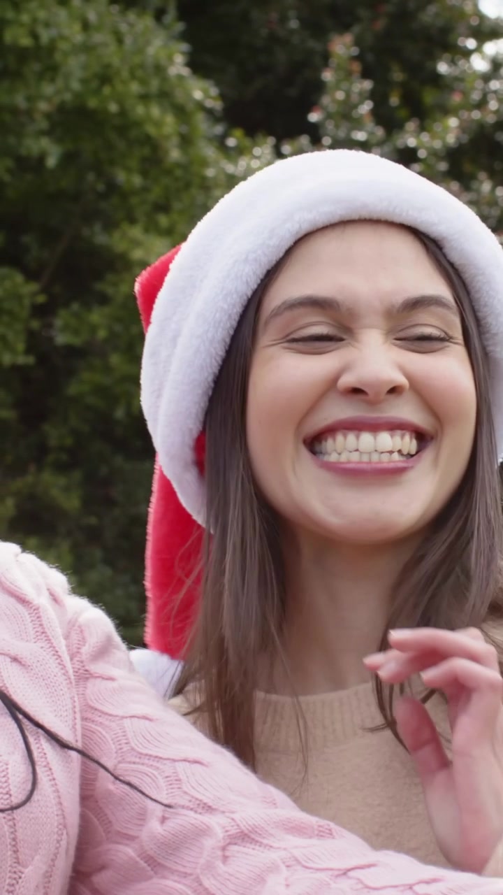 Vertical video: Seeing pink phone, two women in Santa hats and knits laughing and hugging in garden