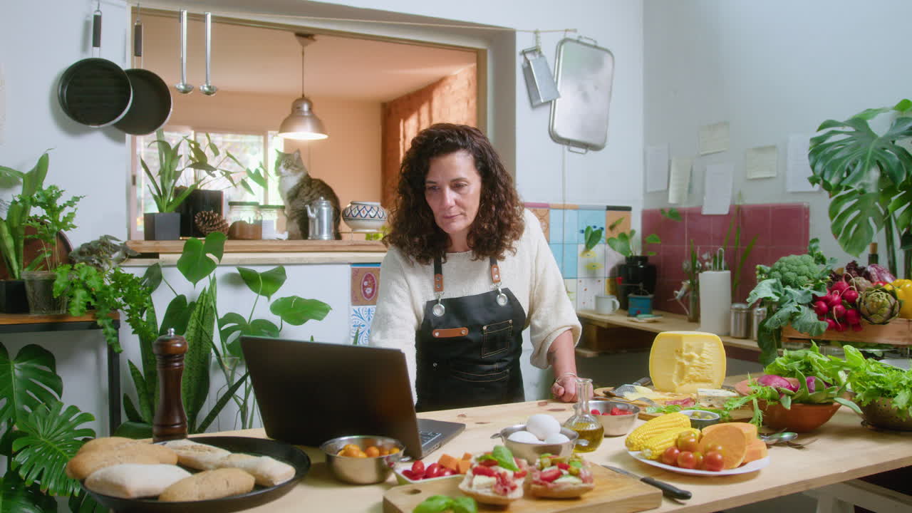 Woman preparing food in the kitchen with a laptop and cat