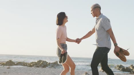 Happy-biracial-couple-walking-and-holding-hands-at-beach,-in-slow-motion