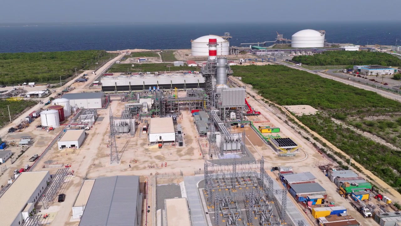 Vista aérea amplia de una gran central eléctrica de gas natural en construcción con infraestructura eléctrica y tanques de almacenamiento cerca de la costa caribeña.