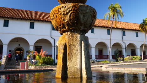 people and tourists walking up the steps of the historic santa barbara mission building with spanish catholic architecture and fountain in the foreground slide left