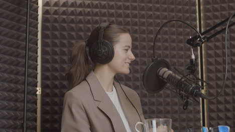 an elegant young woman with her hair tied back and wearing a smiling suit listens smilingly to the person who is talking in a radio recording studio