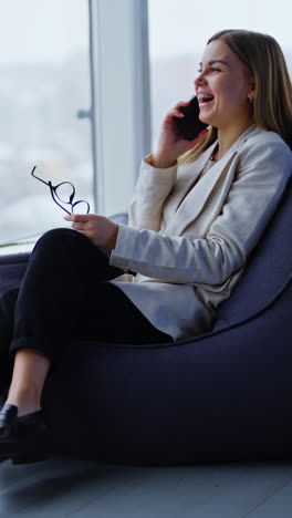 resilient positive lady sits in bean bag chair talking on the phone. woman waves her hand with glasses during conversation. panoramic windows in blur at backdrop. vertical video
