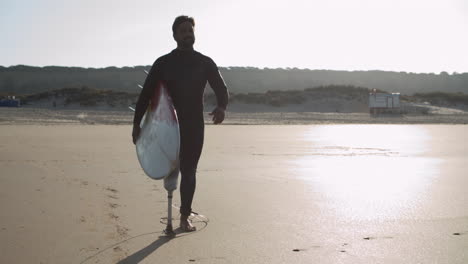 front view of a male surfer with artificial leg walking along beach and holding surfboard under arm