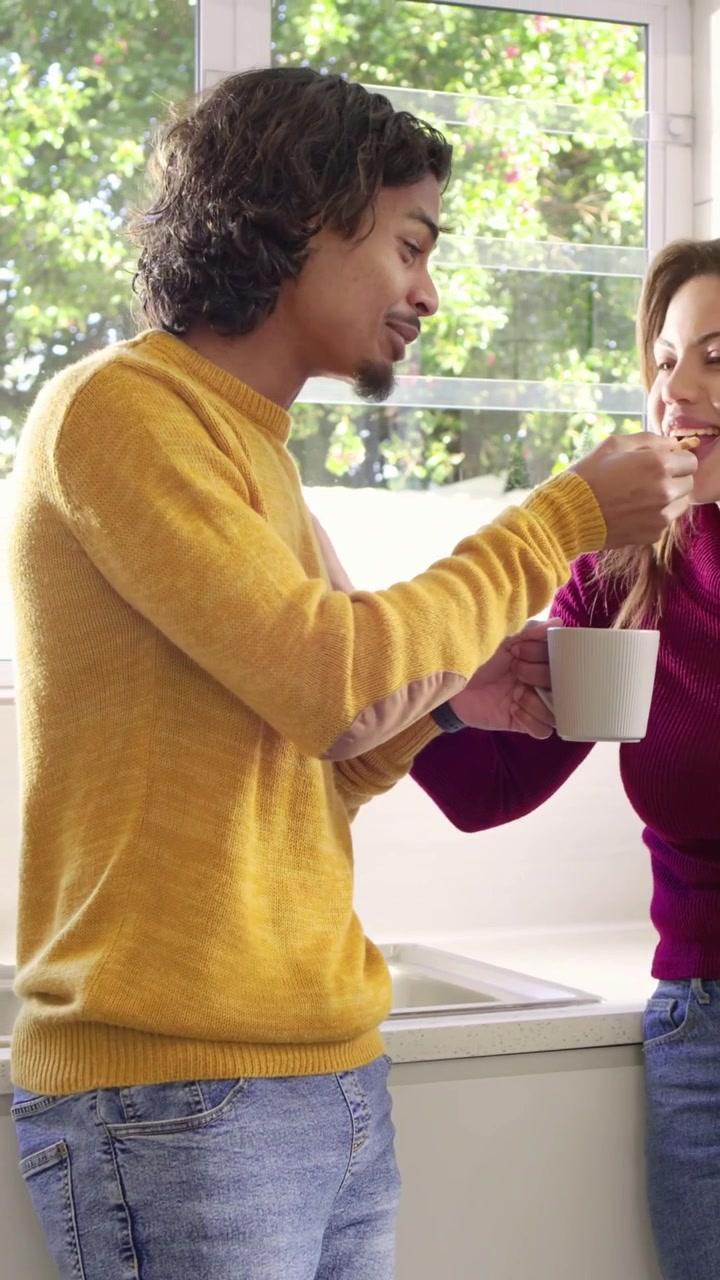 Video verticale: Donna con top color prugna che offre un cucchiaio per assaggiare, uomo con maglione giallo che sorseggia da una tazza in cucina