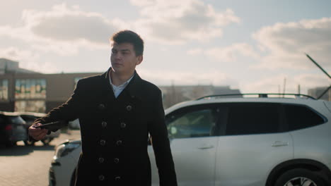 investor in black coat leaving car with serious expression while retrieving phone from pocket to answer call in an urban area featuring parked cars, modern buildings, moving vehicles and greenery