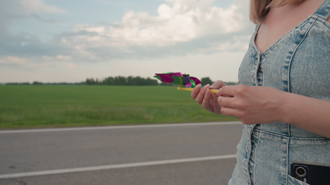 Close up rear view woman walking along country road rotating pinwheel in hand phone visible in denim pocket under cloudy sky beside lush green field in light breeze