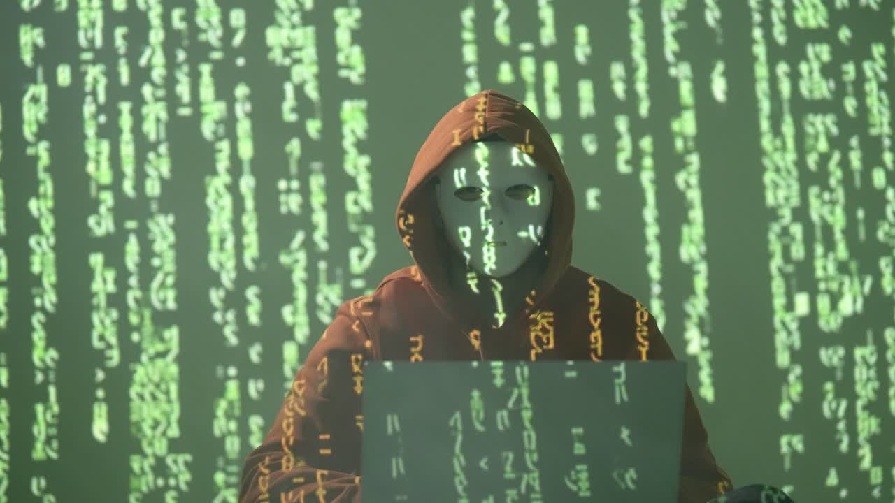 A masked hacker in a hoodie works on a laptop as green code streams down the wall. The scene evokes cybercrime, data hacking, and anonymity in a dark, tech-driven digital environment