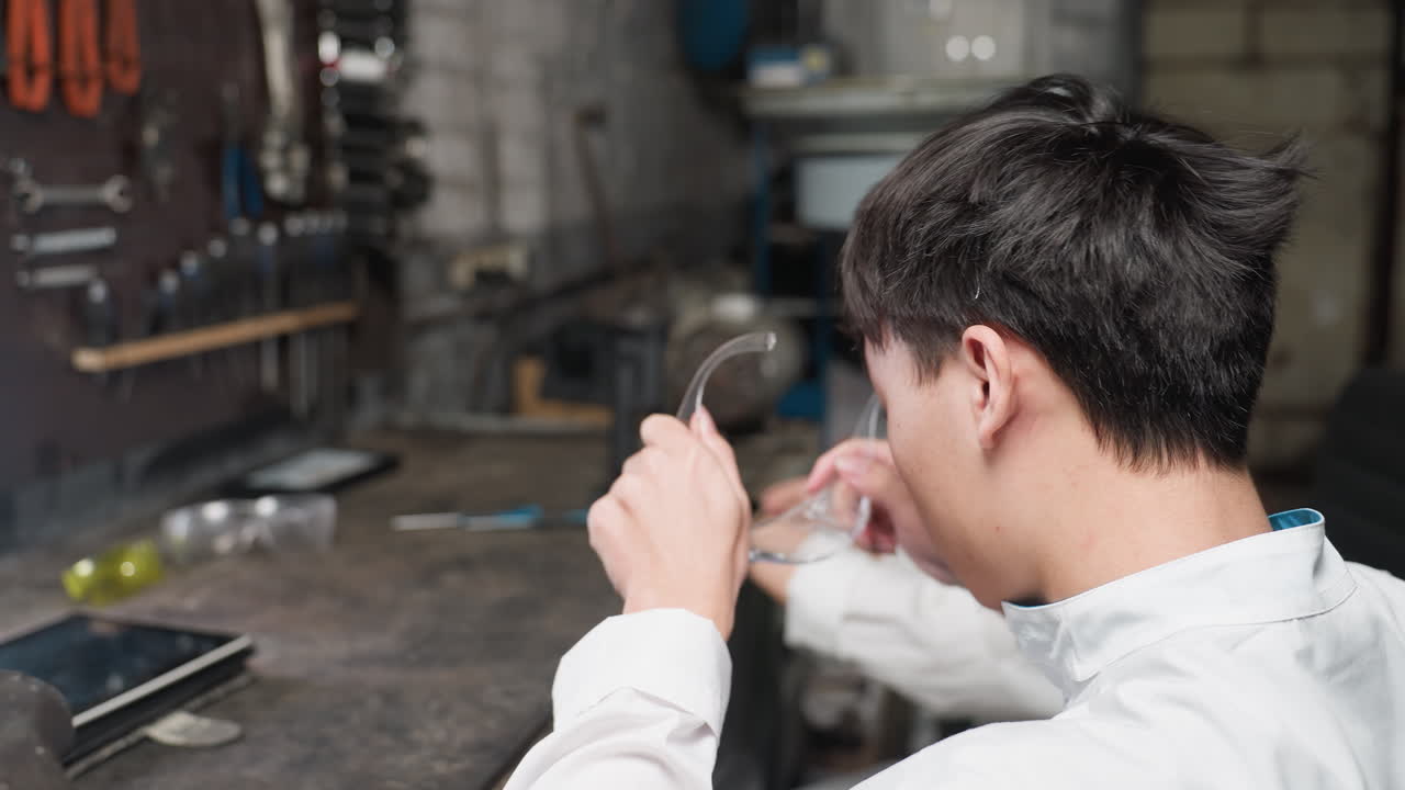 자동차 작업실에 앉아있는 실험실 엔지니어의 클로즈업, 남자는 보호 안경을 착용하고 태블릿을 들고, 여자는 기계와 조직된 장비로 둘러싸인 그녀의 앞에 있는 도구에 초점을 맞추고 있습니다.