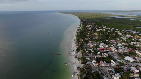 Vista-Aérea-En-Movimiento,-Vista-Panorámica-De-Casas-Y-Edificios-Cerca-De-La-Playa-En-La-Isla-Holbox-En-México,-Pastizales-Y-Cielo-Azul-En-El-Fondo