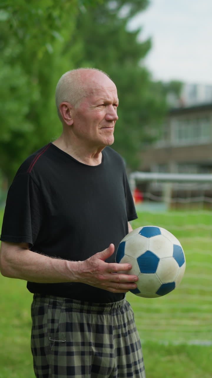 An elderly man rolls a soccer ball in his hand with a focused expression, standing in a grassy field with a building and goalpost in the background