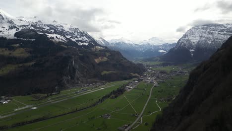 An-aerial-perspective-captures-the-valley-of-Glarus,-Switzerland,-adorned-with-residential-settlements,-embraced-by-the-majestic-snow-capped-peaks-of-the-Swiss-Alps