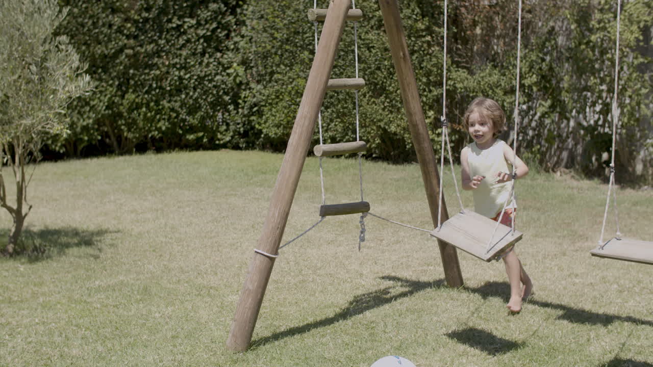 Front view of happy kid in T-shirt and shorts running in the garden, kicking ball and looking at camera