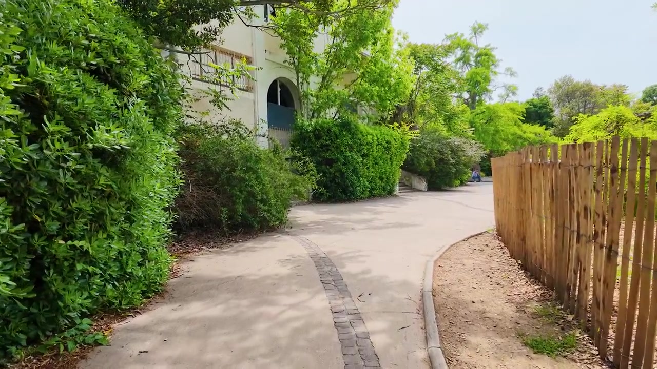 Una telecamera in prima persona si muove in avanti lungo un sentiero lastricato del giardino fiancheggiato da una recinzione di legno.